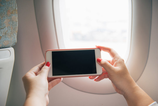 Mockup image of a woman holding and looking at smart phone with blank white screen next to an airplane window with clouds and sky background