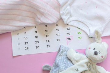 Set of newborn accessories in anticipation of  child - calendar with circled number 26 (twenty six), baby clothes, toys on pink background. Flat lay, top view.