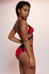 Three quarter side view shot of African lady, wearing bright multicolored swimsuit with high-waisted panties. The woman is posing with hand on waistline, looking to side on light purple background.