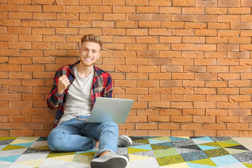 Happy young man with laptop sitting near brick wall