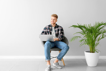 Handsome young man with laptop sitting on chair near light wall