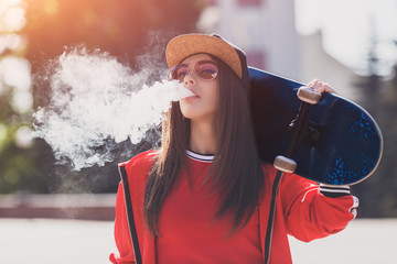 Vaping girl. Young woman with skateboard vape e-cig. Pretty young female in black hat, red clothing vape ecig, vaping device at the sunset. Toned image. Hip-hop style. - 269353001