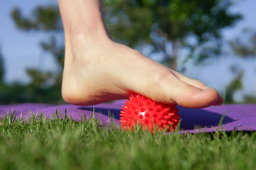 Woman massage feet with spiny plastic red massage ball in summer park. Self myofascial release...