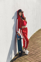 Portrait of young female wearing black hat, red clothing skateboarder holding her skateboard. Woman with skating board looking at camera outdoors.