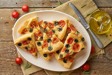 Focaccia, pizza in plate with tomatoes, olives and rosemary. Chopped Italian flat bread, top view, wooden table
