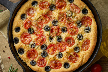 Focaccia, pizza in skillet. Close up italian flat bread with tomatoes, olives and rosemary. Top view, wooden table