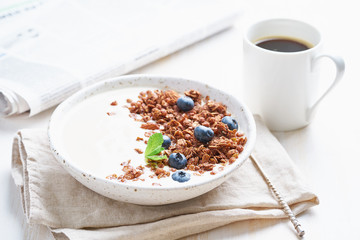 Breakfast with cup of coffee, newspaper, yogurt with chocolate granola, bilberry on a white background, side view