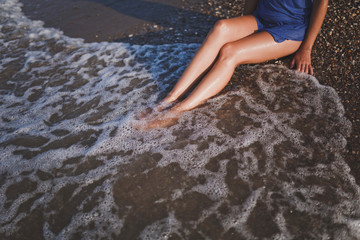 Crop girl with beautiful tanned legs resting in the sea waves on a pebble beach