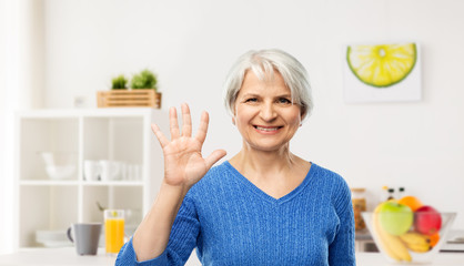 gesture and old people concept - portrait of smiling senior woman in blue sweater showing palm or five fingers over kitchen background