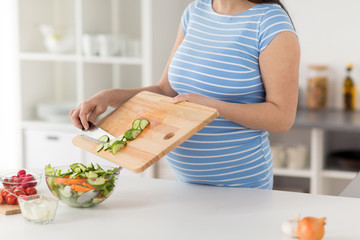 pregnancy, cooking food and healthy eating concept - close up of pregnant woman with kitchen knife adding chopped cucumber from wooden cutting board to salad bowl at home - Powered by Adobe