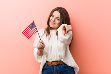 Young curvy plus size woman holding a united states flag cheerful smiles pointing to front.