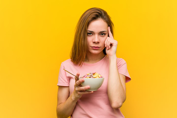 Young woman holding a cereals bowl pointing her temple with finger, thinking, focused on a task.