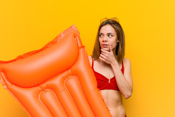 Young woman wearing bikini, holding an air mattress bed looking sideways with doubtful and skeptical expression.