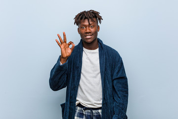 Young african black man wearing pajama winks an eye and holds an okay gesture with hand.