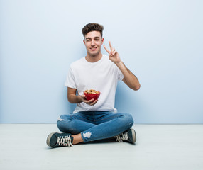 Young man eating cereals sitting on the floor showing victory sign and smiling broadly.