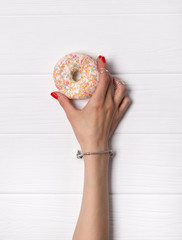  Female hand with red manicure holding donut on white wooden background.