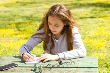 Cute teen girl writes notes on a paper pad by wooden table on a green meadow.