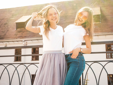 Portrait of two young beautiful blond smiling hipster girls in trendy summer white t-shirt clothes. Sexy carefree women posing on street background. Positive models having fun in sunglasses