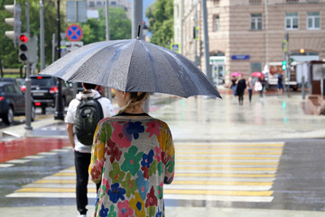 Rainy day in a city, girl with umbrella standing on a street near the pedestrian crossing. Summer rain, weather concept