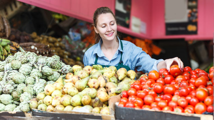 seller is standing near shelves with vegetables and fruits
