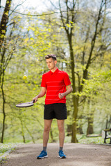 The man in a red t-shirt pose with a tennis racket and a ball on the background of green park. Sport concept