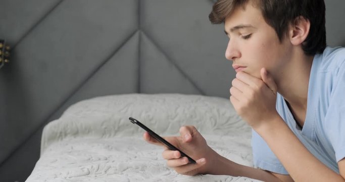 Teenager upset by the bad news. Teenage boy is shocked by a bad sms message. White young man depressed and hides his face. Caucasian student lying on a bed with cell phone. Real time