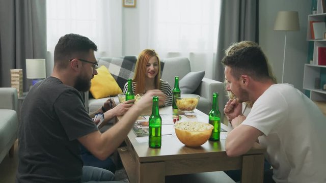 Company of boys and girls playing board game in living room and drinking beer