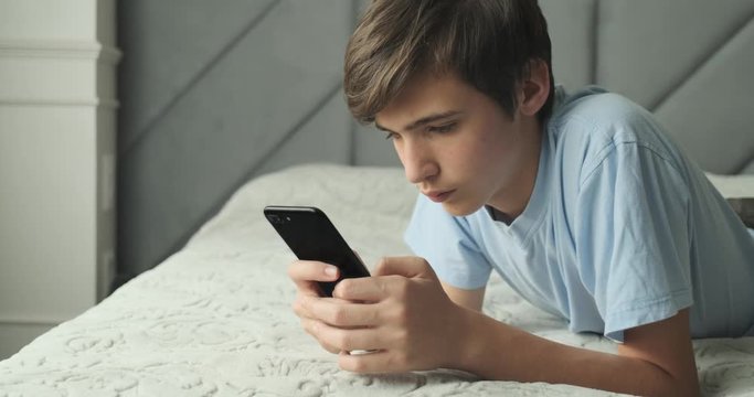 Teenager typing text message by smart phone, at home. Handsome boy spending time in a social network using mobile phone lying on sofa. Caucasian teenage guy talking on cell phone. Lifestyle concept.