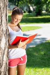 young brunette woman reading to notepad, leaning on a tree