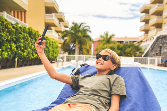 Young boy relaxing outdoor and taking selfie with smartphone. Trendy child get connected on line with remote friends near a swimming pool after school. Technology allows to keep in touch everywhere