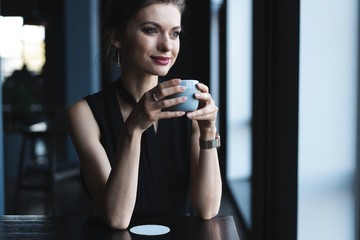 Portrait of gorgeous female drinking tea or coffee and looking with smile out of the coffee shop window while enjoying her leisure time, nice business woman lunch in modern cafe during her work break.