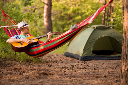 young caucasian man in hat play guitar and swinging in hammock of weekend morning in camp rest