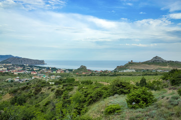 Fototapeta na wymiar Sudak, Crimean Republic, Russia - May 30, 2017: View of the district of Sudak, residential buildings. Terrain in the mountains, vineyards, the sea on the horizon. View of Alchak mountain.
