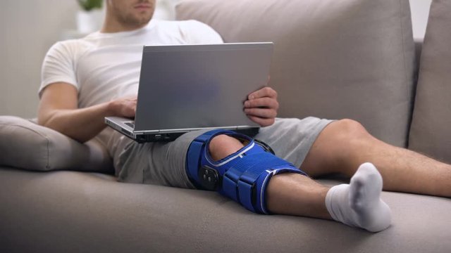 Man in arthritis knee brace working on laptop at home rehab period and freelance