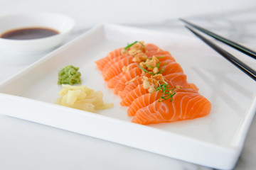 Sliced salmon sashimi, a popular Japanese food, on white plate with wasabi, pickled ginger and soy sauce on the side shown with chopsticks and white background