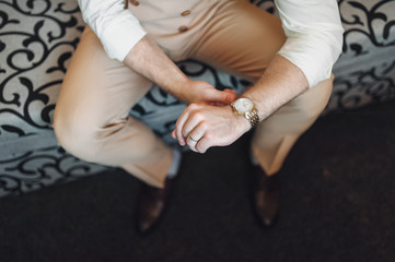The groom in a beige suit and waistcoat sits on the couch and straightens the clock. Waiting for...