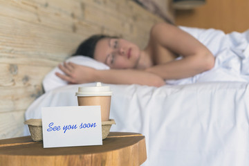A paper cup of coffee and sticker with phrase "see you soon" standing on the table in the bedroom as a surprise for a sleeping girl.