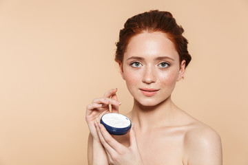 Amazing young redhead woman posing isolated over beige wall background take care of her skin with cream.