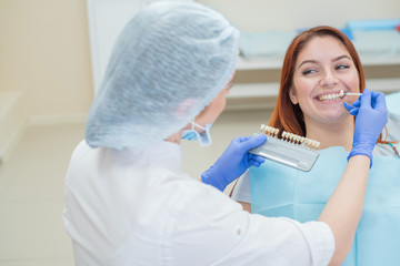 Naklejka premium Check and select the color of the teeth in the dentist chair. Dentist makes the process of treating a beautiful young red-haired girl. Selection of a tooth implant.