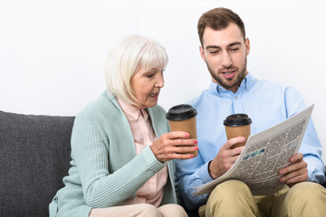 man and senior mother holding cups of coffee and reading newspaper at home