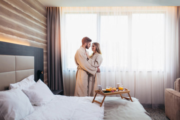 Young happy couple in hotel room in the morning. Just married man and woman standing at window. - obrazy, fototapety, plakaty