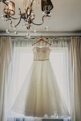 Wedding dress. White wedding dress with a full skirt on a hanger in the room of the bride with white curtains. Wedding attributes. No people.