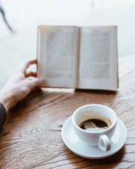 Black coffee on saucer with Human hand holding vintage book on blurred background on the wooden table next to big coffee shop window.