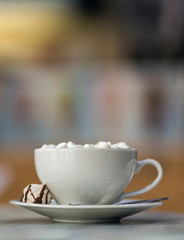 Mug of coffee with marshmallows on porcelain plate on blurred colorful interior bokeh background.