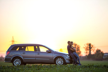 Young couple, slim attractive woman with long ponytail and handsome man standing at silver car in green field on clear sky at sunset or sunrise copy space background.