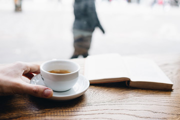 Human hand holding black coffee on saucer with vintage book on background on the wooden table next to big coffee shop window and looking to passerby street walkers