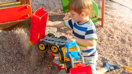 Portrait of cheerful smiling little boy pouring sand in toy truck with trailer. Kids playing and...
