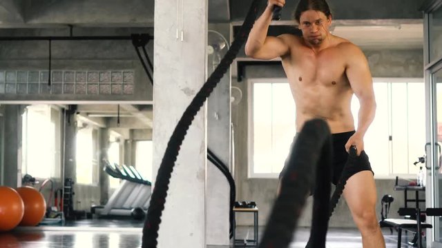 Muscular powerful young man training with battle rope doing exercise in functional training fitness gym. Caucasian male fit model working out indoors