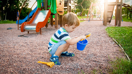 Portrait of little 3 years old toddler boy sitting on the playground and digging sand with toy plastic spade