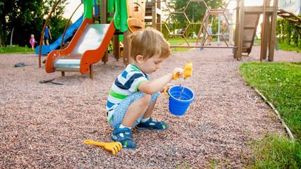 Photo of adorable 3 years old little boy sitting on the playground and digging sand with small plastic shovel and bucket